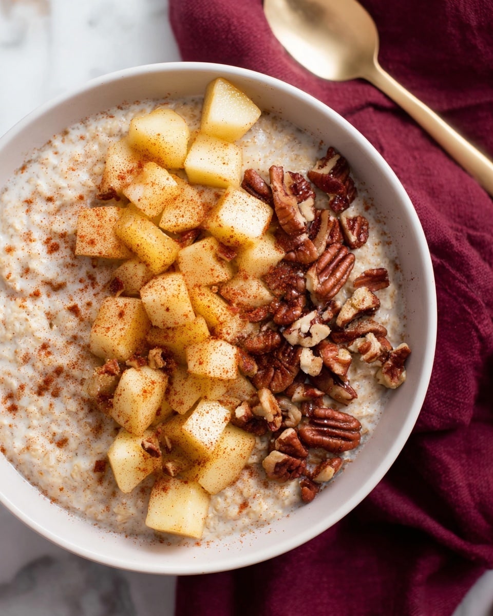 A white bowl filled with a creamy oatmeal base, topped in two sections: one side with small chopped pieces of cooked apples coated with cinnamon, showing shades of light yellow and warm brown, and the other side with roughly chopped pecans in varying browns and cream, all placed on a white marbled surface with a folded deep maroon cloth nearby and a golden spoon partially visible in the corner, photo taken with an iphone --ar 4:5 --v 7