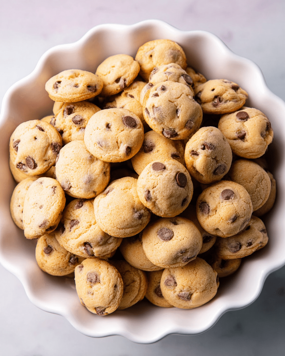 A white scalloped-edged bowl filled with many small, round chocolate chip cookies that are golden brown with darker chocolate chips scattered throughout. The cookies are piled in multiple layers, showing their soft texture and some lightly browned undersides. The bowl sits on a surface with a white marbled texture. photo taken with an iphone --ar 4:5 --v 7