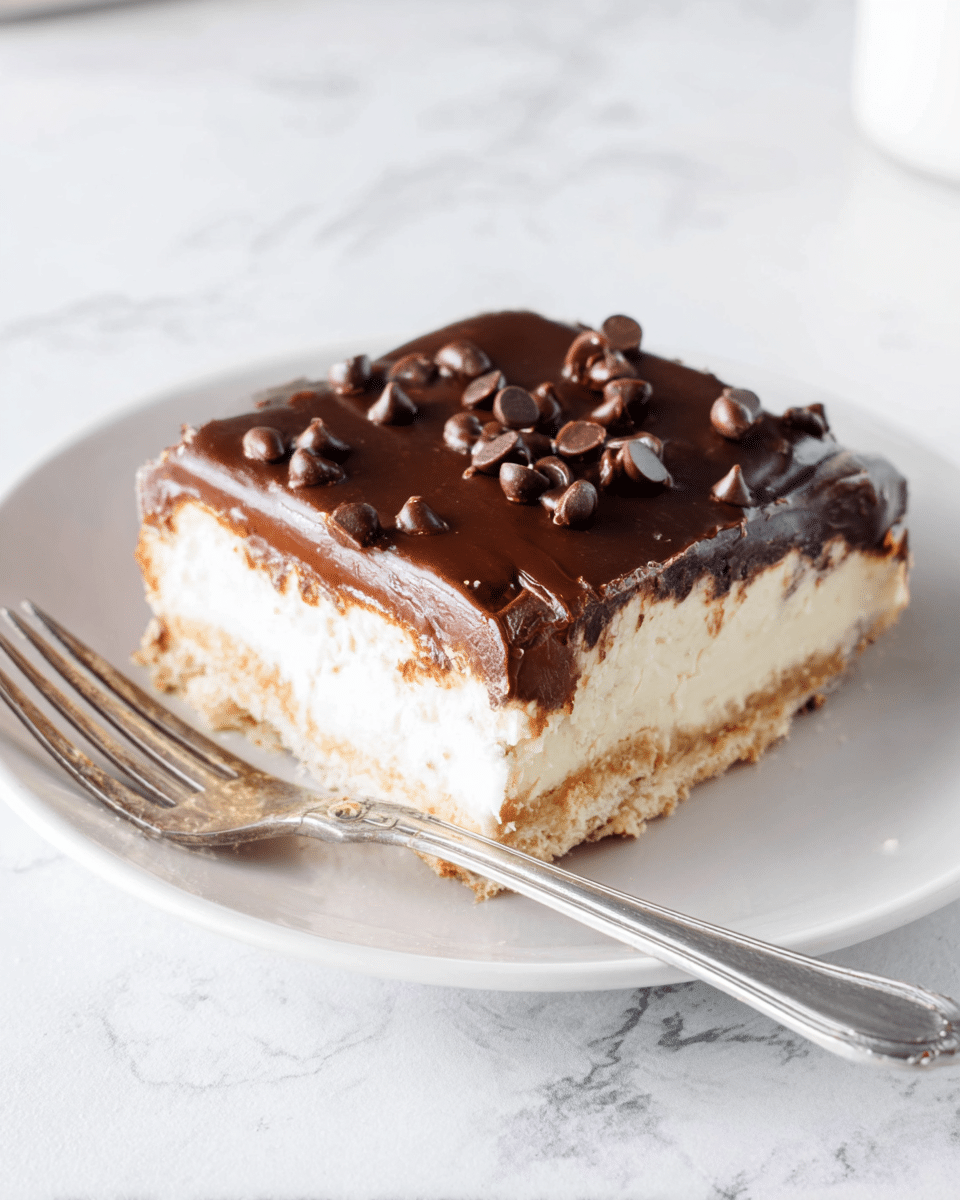 A square slice of dessert with three clear layers sits on a white plate. The bottom layer is a light golden crust with a slightly crumbly texture. The middle layer is thick and creamy, off-white in color, smooth and dense. The top layer is a glossy, dark chocolate coating scattered with small, shiny chocolate chips. A silver fork rests on the plate beside the dessert, and the background shows a white marbled surface. photo taken with an iphone --ar 4:5 --v 7