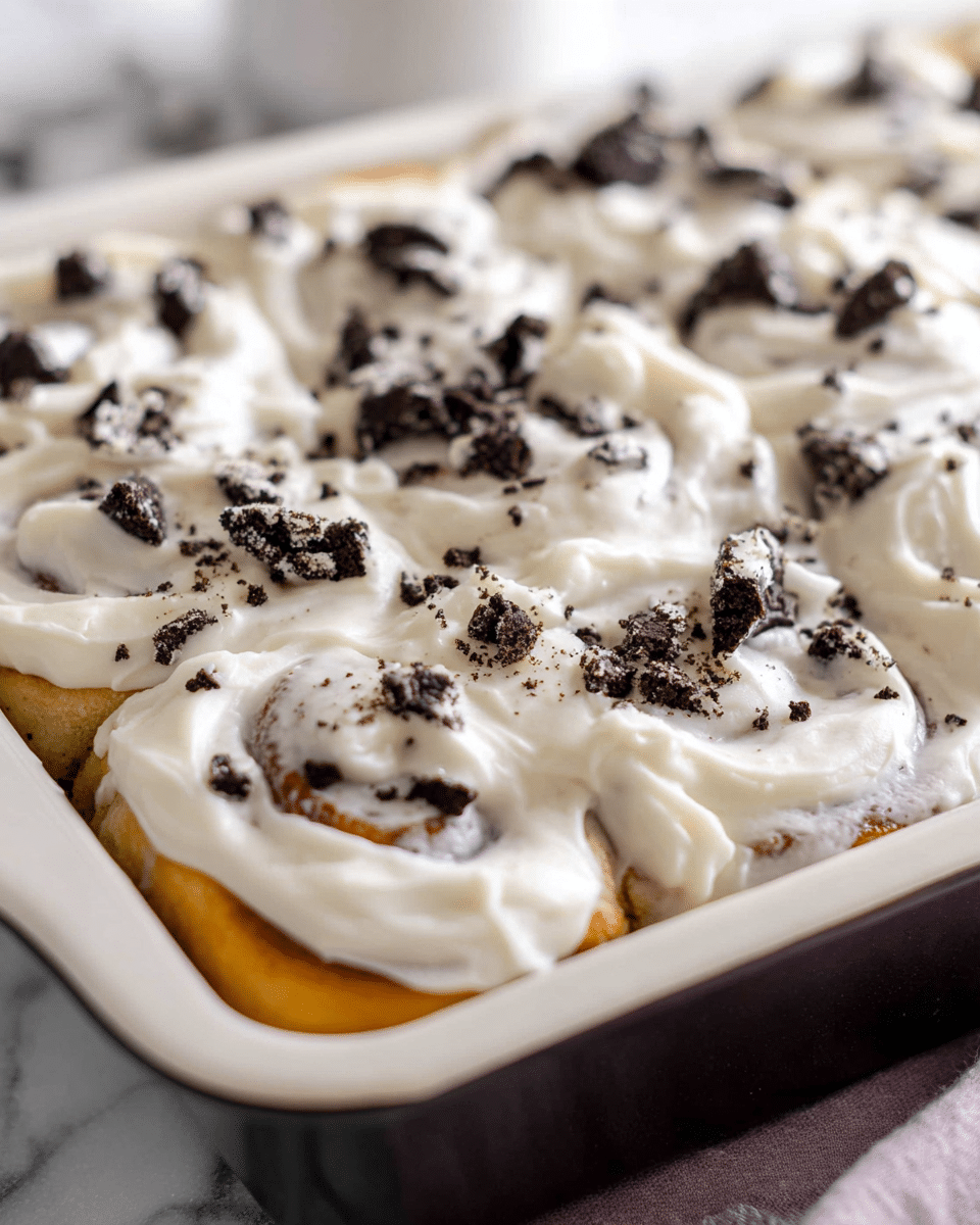 The image shows a close-up of a baking pan filled with soft cinnamon rolls covered in a thick, creamy white frosting. On top of the frosting, there are small pieces of crushed black and white cookies scattered all over, adding a textured look. The cinnamon rolls themselves are golden brown and swirled underneath the smooth layer of frosting. The pan is resting on a white marbled surface. photo taken with an iphone --ar 4:5 --v 7