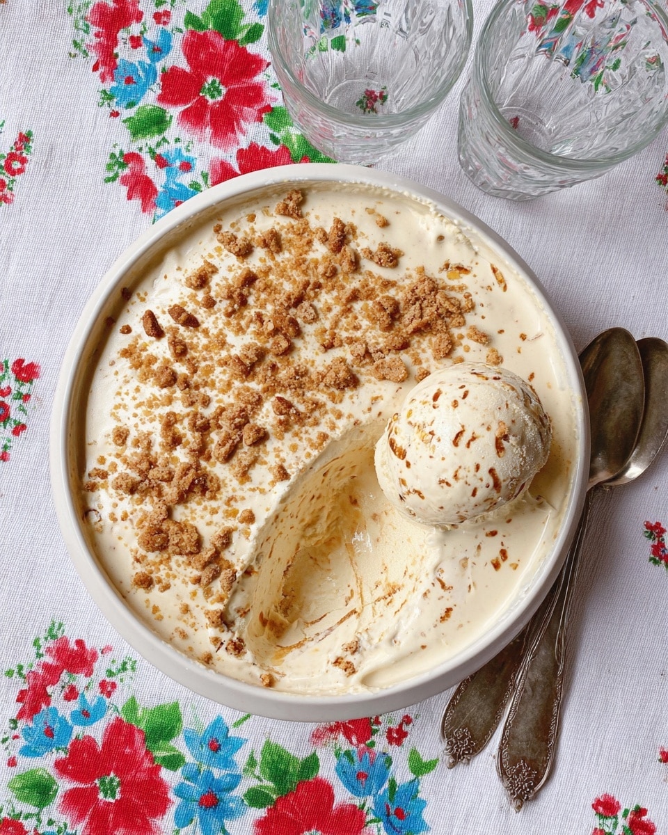 A round white bowl contains a frozen dessert that appears light cream-colored with crumbled brown biscuit pieces sprinkled thickly on top and throughout the inside; one scoop of the dessert, speckled with cookie bits, rests on the right edge where a section has been scooped out, revealing a creamy, textured interior of the same color and brown crumbs. The bowl is set on a white cloth decorated with a colorful flower pattern featuring red, blue, and green hues. Two tarnished silver spoons lie below the bowl, while two clear glass bowls are placed above it on the cloth. photo taken with an iphone --ar 4:5 --v 7
