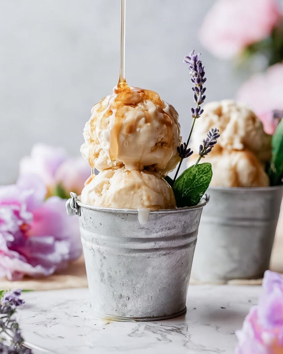 The image shows two small metal buckets on a white marbled surface. Each bucket holds two large scoops of creamy beige ice cream with a smooth, slightly melting texture. Golden syrup is being poured over the front bucket’s ice cream, creating a shiny drizzle. A green leaf and delicate purple lavender sprigs stick out from the ice cream in the front bucket. The background is softly blurred with pastel pink flowers and more lavender, creating a calm, fresh look. Photo taken with an iphone --ar 4:5 --v 7