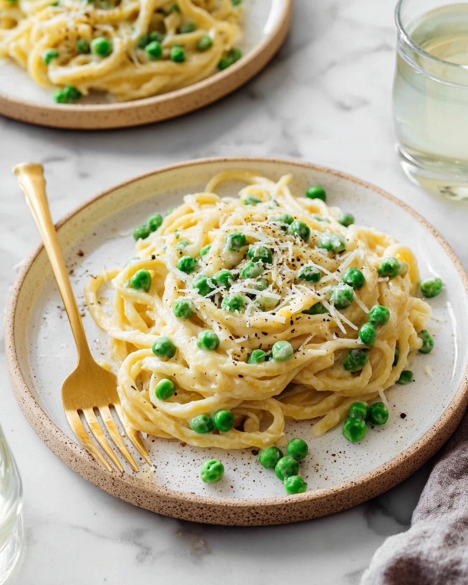 A white speckled plate holds a serving of thick yellow pasta noodles, topped with bright green peas scattered evenly on top and mixed within. A creamy white sauce coats the noodles, with fine shreds of pale yellow cheese sprinkled over them. Light black pepper specks add a subtle contrast. A golden fork rests on the left side of the plate, partially placed on the pasta. In the background, there is another similar plate of pasta, all set on a white marbled surface. A clear glass of water is partially visible to the right. photo taken with an iphone --ar 4:5 --v 7