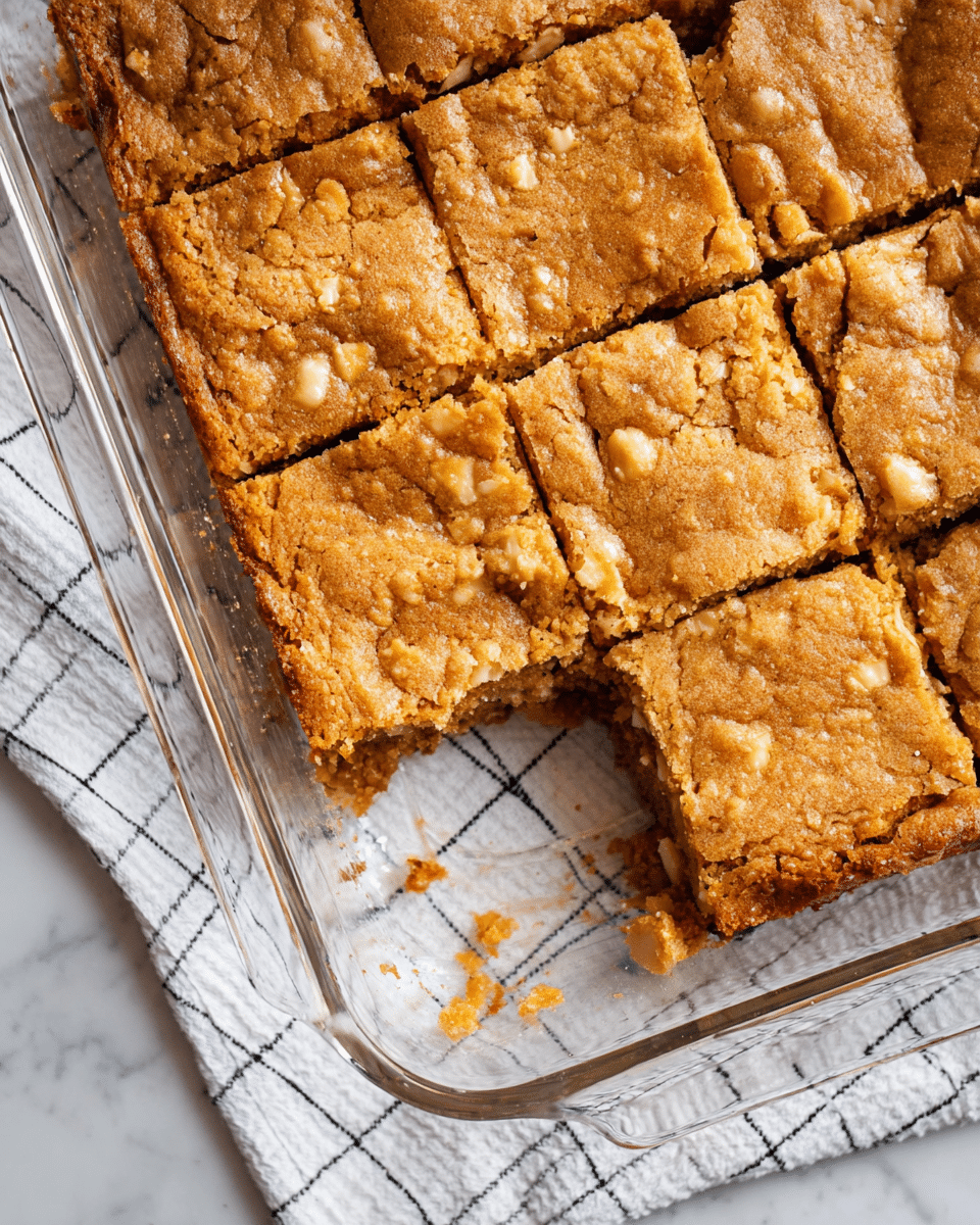 A glass baking dish with nine square pieces of blondies, one piece missing from the bottom right corner, showing a thick, golden brown, crumbly top layer with a slightly cracked texture. Beneath the top crust, the blondies have a moist, dense, and chewy interior with visible white chocolate or nut bits scattered throughout. The dish is placed on a white marble surface covered by a white cloth with a black grid pattern. photo taken with an iphone --ar 4:5 --v 7