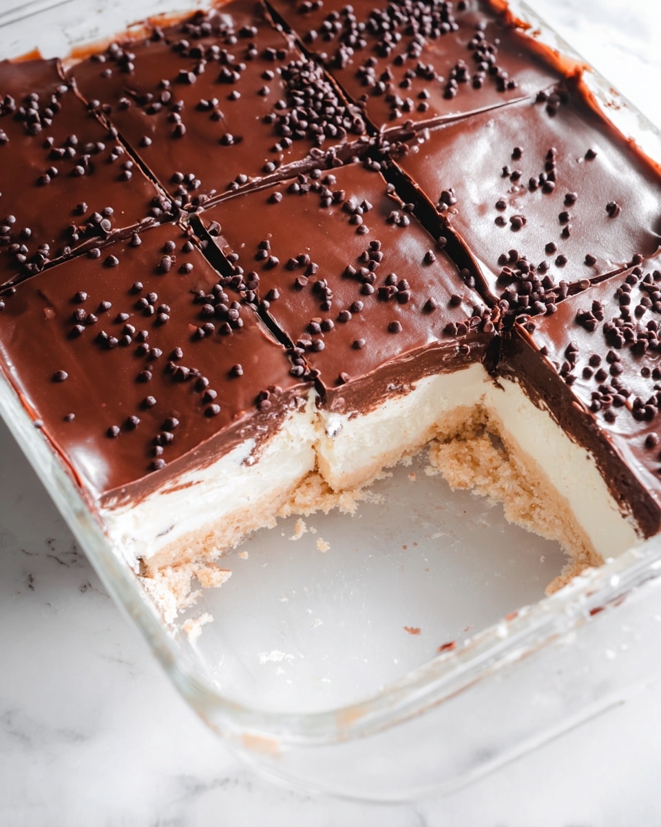 The image shows a dessert cut into square pieces inside a clear glass dish. The dessert has three layers: the bottom layer is pale beige and crumbly, the middle layer is creamy white and smooth, and the top layer is a thick, shiny dark brown chocolate ganache sprinkled with small chocolate chips. A few pieces have been removed from the dish, revealing the layers clearly. The dish sits on a white marbled surface. photo taken with an iphone --ar 4:5 --v 7