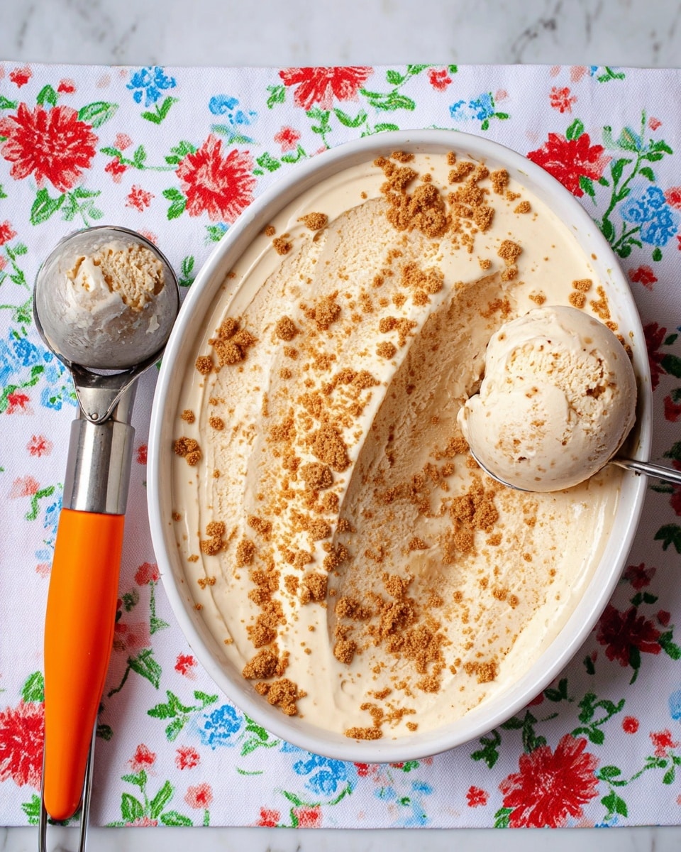 The image shows a white round dish filled with creamy ice cream that has a light beige color and is topped with small brown crumb pieces scattered evenly across the surface. A scoop has been taken out from the ice cream on the right side, revealing its smooth texture and the brown crumbs inside. The scoop of ice cream rests near the empty part of the dish and has the same beige color with brown speckles. Next to the dish is an ice cream scoop with a metal head and bright orange handle. The scene is set on a white marbled tablecloth with colorful floral patterns in red, blue, and green. Photo taken with an iphone --ar 4:5 --v 7