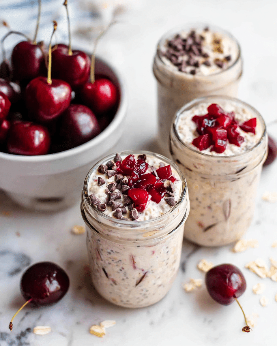 The image shows three glass jars filled with a creamy, oat-based mixture that appears light beige with visible oats and small dark bits, likely chia seeds or chocolate chips, giving the texture a speckled look. The top layer in each jar is decorated with two sections: one side has small dark chocolate chips, clustered neatly, and the other side is garnished with chopped, bright red cherries. Behind the jars, there is a white bowl filled with whole, dark red cherries with stems, creating a fresh and colorful backdrop. The jars and bowl sit on a white marbled surface with some scattered oat flakes and a few chocolate chips around them. Photo taken with an iphone --ar 4:5 --v 7