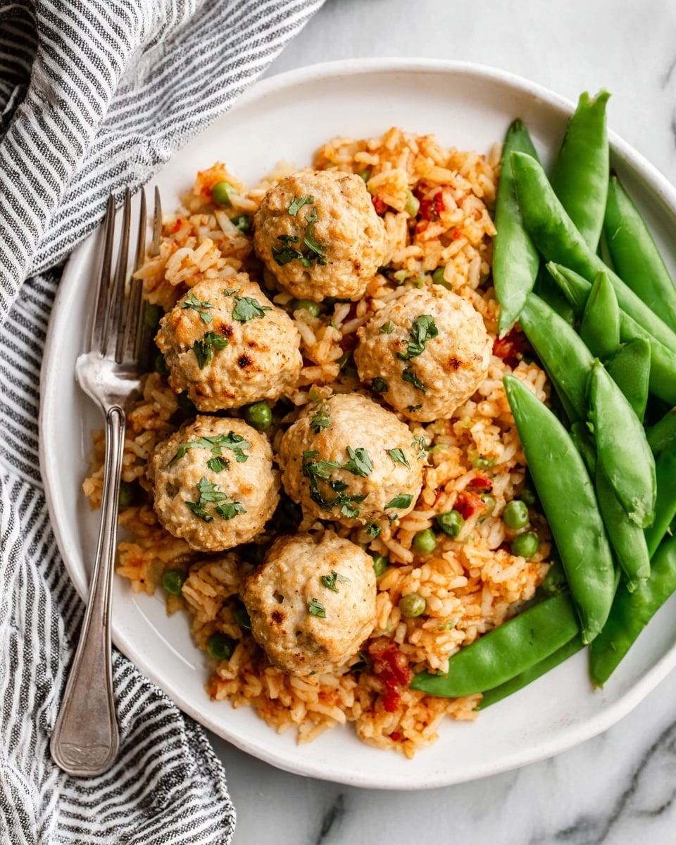 A white plate on a white marbled surface holds a colorful meal with three main layers: at the bottom, there is a bed of mixed rice that looks fluffy and has orange, red, and green bits mixed in, giving it a textured look; on top of the rice, there are eight light brown meatballs evenly placed, each with small herbs visible on the surface, adding green specks; to the right side of the plate, a neat pile of bright green snap peas adds a fresh, smooth texture and color contrast. A silver fork rests on the left edge of the plate, and a striped cloth napkin sits beneath it. The photo taken with an iphone --ar 4:5 --v 7