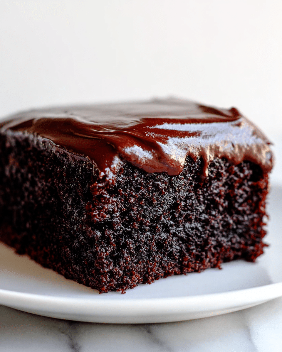 A close-up view of a single square piece of chocolate cake on a white plate, showing two layers: a thick, dark, moist and crumbly chocolate cake base, topped with a shiny, smooth, dark chocolate frosting that slightly drips over the edges. The cake sits on a white marbled surface with a plain light background. Photo taken with an iphone --ar 4:5 --v 7