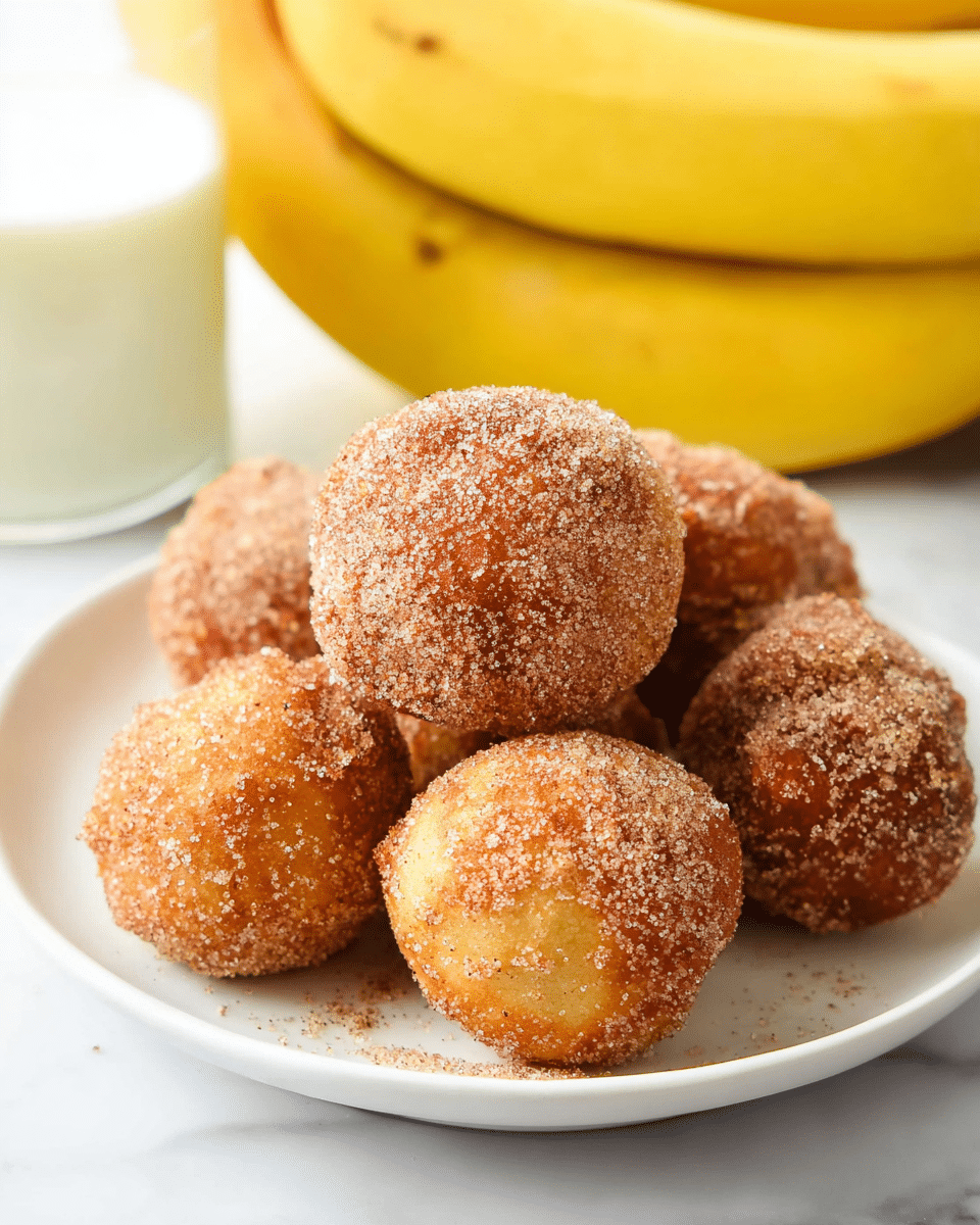 A white plate holds a small pile of round, golden-brown donut holes covered in a grainy layer of cinnamon sugar. The donut holes appear fluffy and soft, with a slightly rough sugar texture on the outside. In the background, there is a bunch of ripe yellow bananas and a glass of milk, all set on a white marbled surface. The image focuses closely on the donut holes, showing their detailed sugary coating and warm color, suggesting a fresh, warm treat. photo taken with an iphone --ar 4:5 --v 7