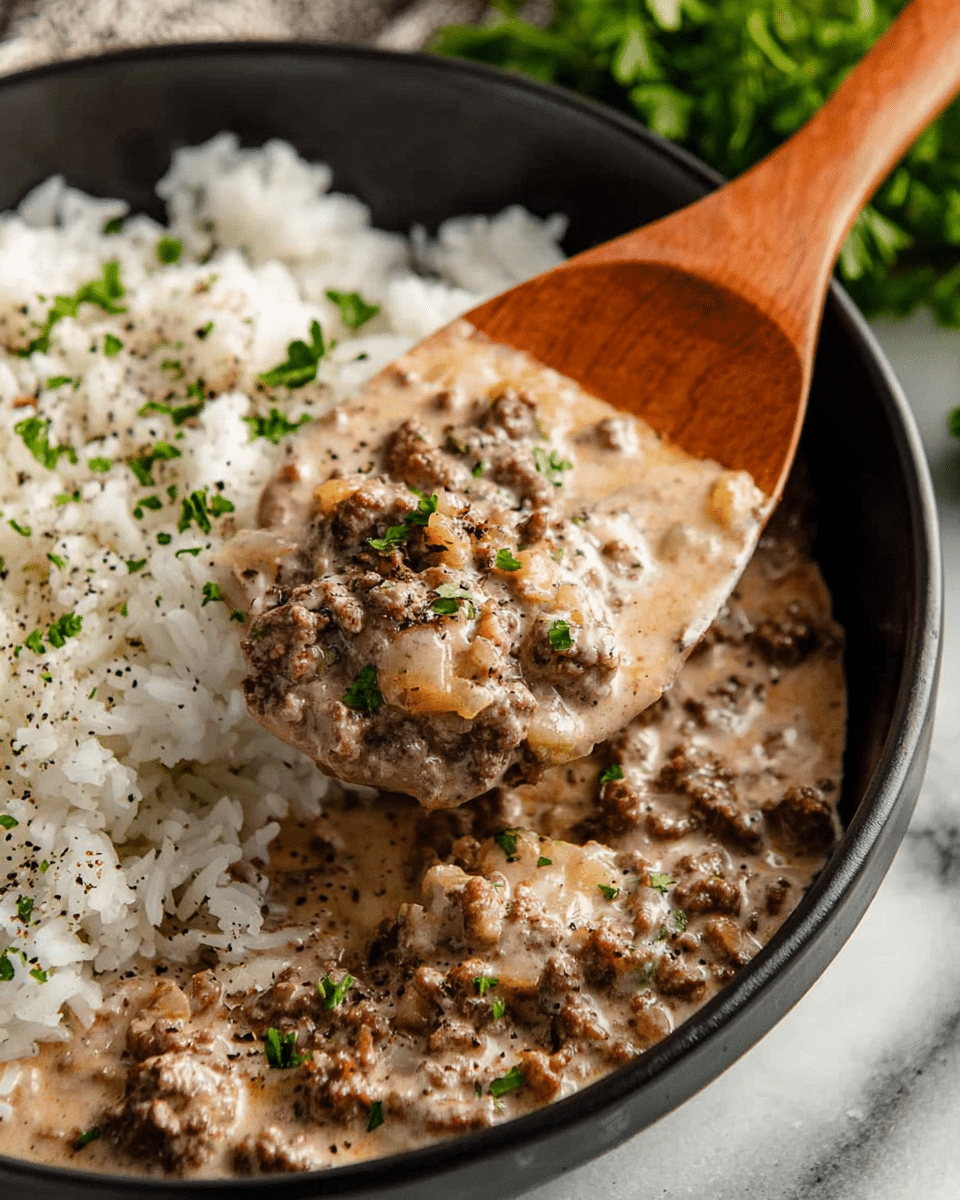 A close-up of a black bowl filled halfway with white rice sprinkled with chopped green herbs and black pepper, next to a creamy mixture of ground beef and diced onions in a light brown sauce covering the other half of the bowl; a wooden spatula lifts a portion of the creamy beef mixture showing its thick texture, all set on a white marbled surface with some green herbs blurred in the background. Photo taken with an iphone --ar 4:5 --v 7