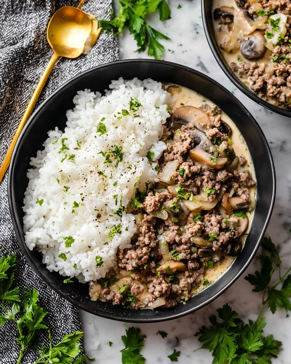 A black bowl holds two main layers: on one side, white rice with a fluffy texture sprinkled lightly with chopped green herbs and black pepper, and on the other side, a creamy mixture of browned ground beef, sliced mushrooms, and translucent cooked onions coated in a light beige sauce speckled with green herbs. The dish sits on a white marbled surface scattered with fresh green parsley leaves. A gold spoon is beside the bowl, and a textured gray cloth adds contrast nearby. Another similar bowl is partially visible in the top right background. Photo taken with an iphone --ar 4:5 --v 7