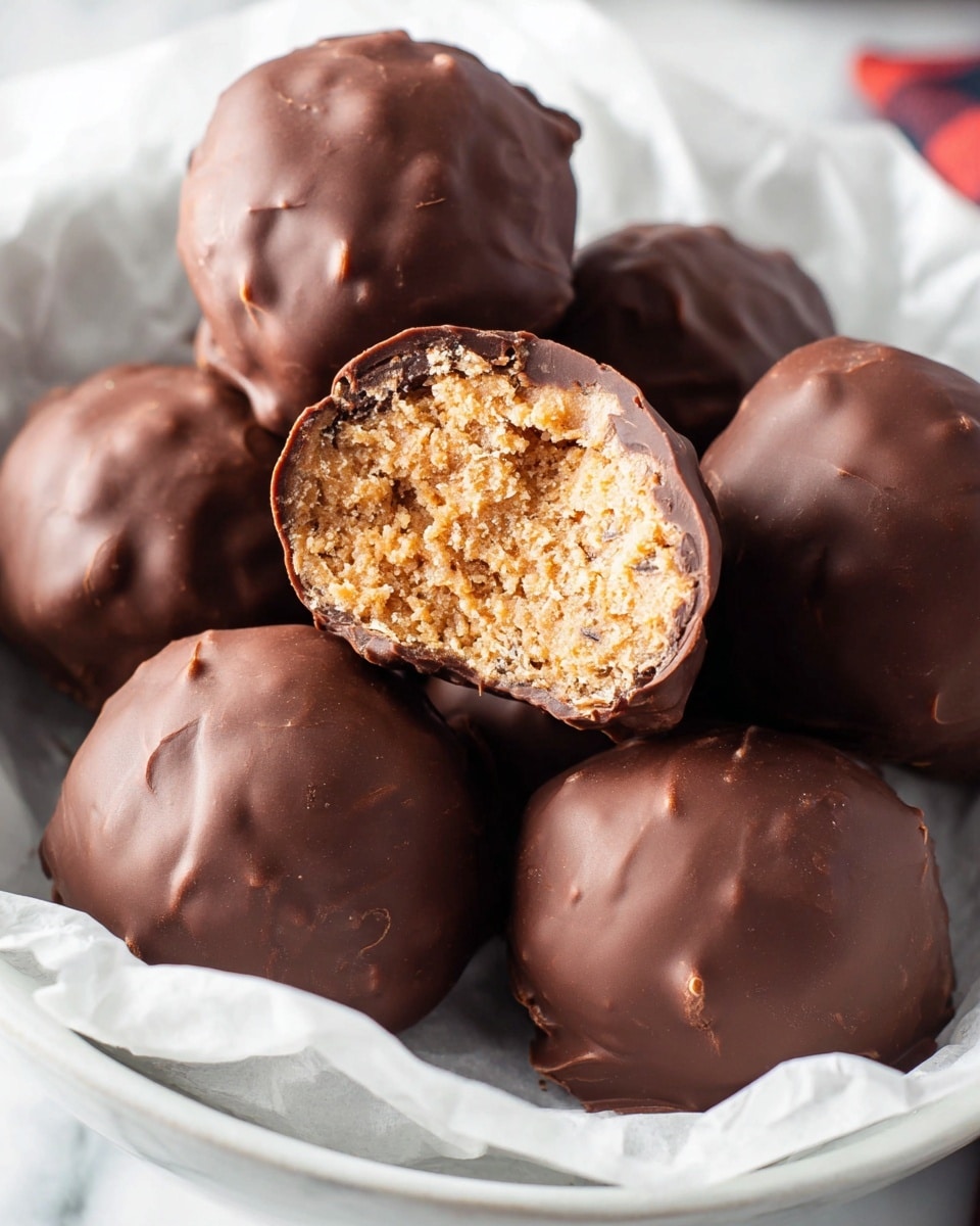 The image shows six round chocolate-covered treats placed in a bowl lined with white parchment paper, set on a white marbled surface. One treat in the center is cut in half, revealing a light brown, crumbly, and textured filling inside with slight darker brown streaks near the chocolate shell. The outer layer is a smooth, dark chocolate coating with a glossy finish and some small wrinkles. The treats have an uneven, hand-formed shape with occasional dimples and bumps on the surface. photo taken with an iphone --ar 4:5 --v 7