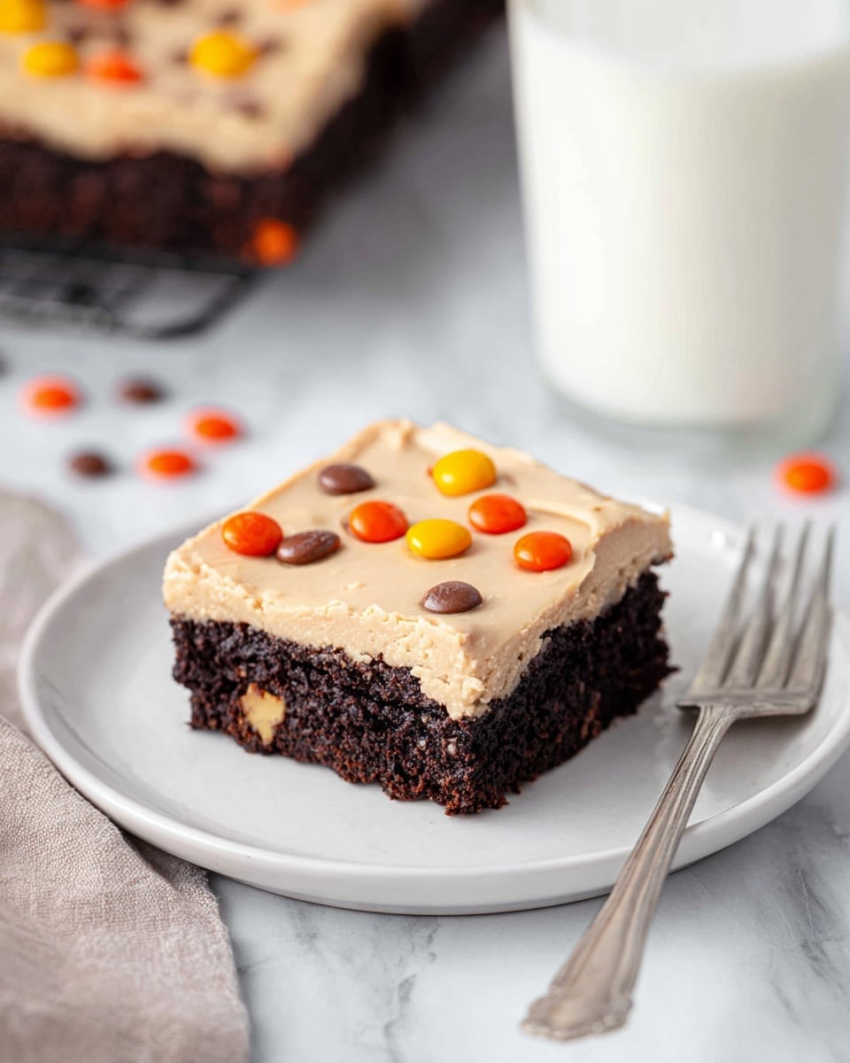 A single square brownie sits on a white plate placed on a white marbled surface. The brownie has two layers: a thick, dense, dark brown chocolate base with a moist texture, and a smooth, creamy light tan frosting spread evenly on top. Small pieces of orange, yellow, and brown candy-coated chocolates are scattered on the frosting, adding bright spots of color. In the background, a glass of milk and part of a second brownie on a cooling rack are softly out of focus. A silver fork lies near the bottom right of the plate. photo taken with an iphone --ar 4:5 --v 7