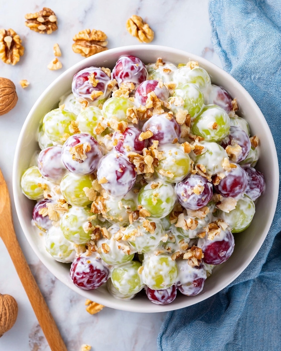 A white bowl filled with two layers of grapes covered in a thick, creamy white dressing; the grapes are green and red, mixed evenly throughout. The top layer is sprinkled with small pieces of chopped walnuts, adding a light brown, crunchy texture. The bowl sits on a white marbled surface with scattered walnuts and grapes nearby, and a soft blue cloth partially visible to the right. Photo taken with an iphone --ar 4:5 --v 7