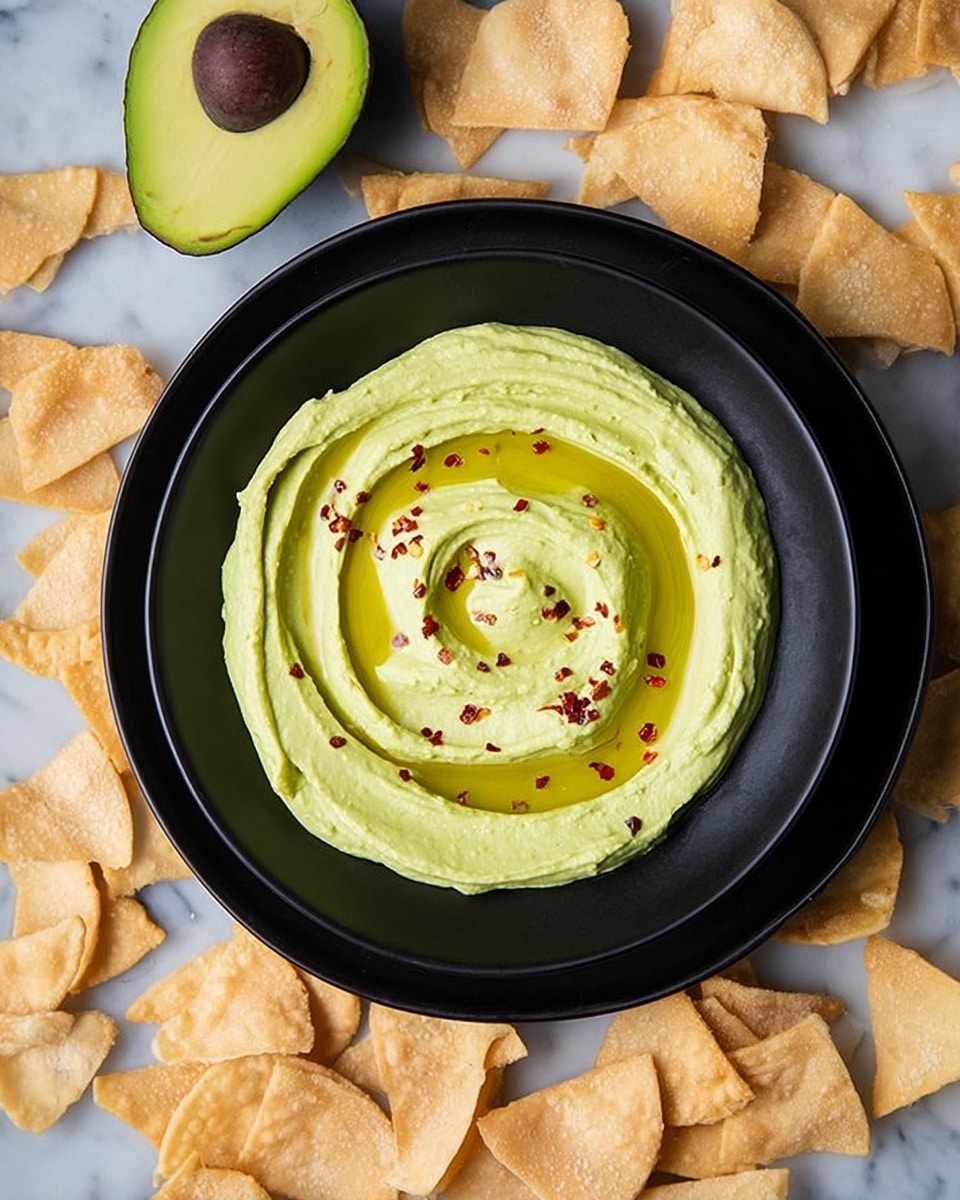 A black bowl filled with one layer of smooth light green avocado hummus, swirled in a circular pattern and topped with a pool of golden olive oil and small red pepper flakes scattered on top. The black bowl is placed on a larger black plate, and both sit on a white marbled surface scattered with many pieces of light golden pita chips. Two halves of a ripe avocado with a dark brown seed and bright green flesh are visible near the top left corner. Photo taken with an iphone --ar 4:5 --v 7