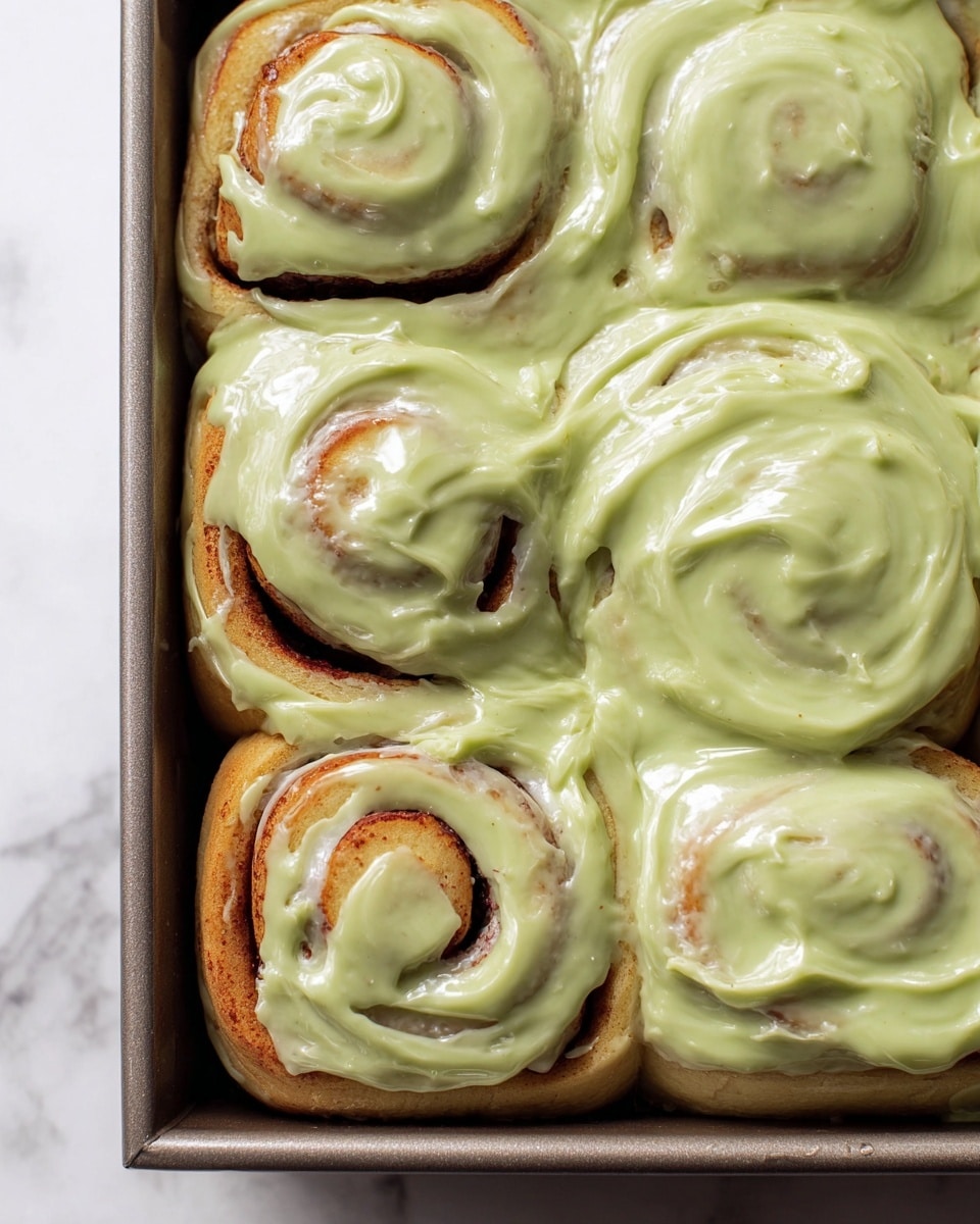 The image shows six cinnamon rolls in a baking pan, each covered with a thick, glossy light green icing that looks creamy and smooth. The cinnamon rolls have a soft, golden-brown color underneath the icing, visible in the swirls and layers of the dough. The rolls are tightly packed, and the icing spreads unevenly but generously over the tops and gaps between the rolls, giving a rich, melting effect. The pan rests on a white marbled surface, adding a clean and fresh background to the close-up shot. photo taken with an iphone --ar 4:5 --v 7