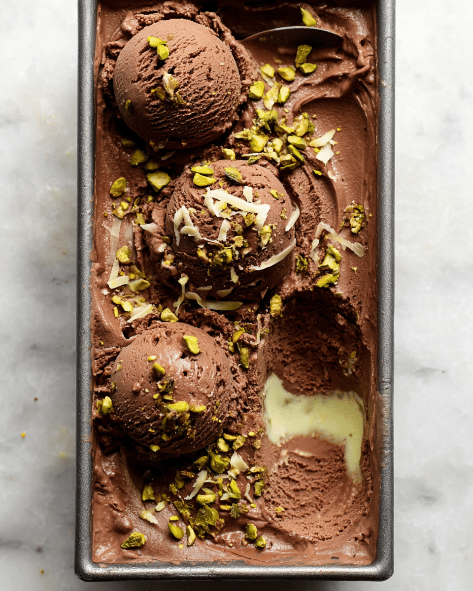 The image shows a rectangular metal container filled with smooth, creamy chocolate ice cream layered evenly inside. On the surface, there are three scoops of rich, dark brown chocolate ice cream, with small bits of crushed green pistachios and thin light beige coconut shreds sprinkled on top. The ice cream texture looks soft and dense with some small air pockets visible. One side of the container has a scoop missing, revealing a pale green layer beneath the chocolate. The container is set on a white marbled surface. photo taken with an iphone --ar 4:5 --v 7
