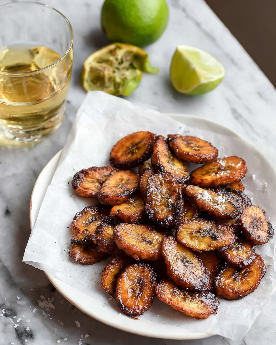 A white plate holds a layer of white parchment paper topped with around twenty roasted plantain slices. Each plantain piece is dark golden brown with some black charred edges, showing a crispy texture with sea salt flakes sprinkled on top. In the background, a glass of light amber liquid sits to the left, while a squeezed lime half and a whole lime with some peeled green skin sit on a white marbled surface. photo taken with an iphone --ar 4:5 --v 7