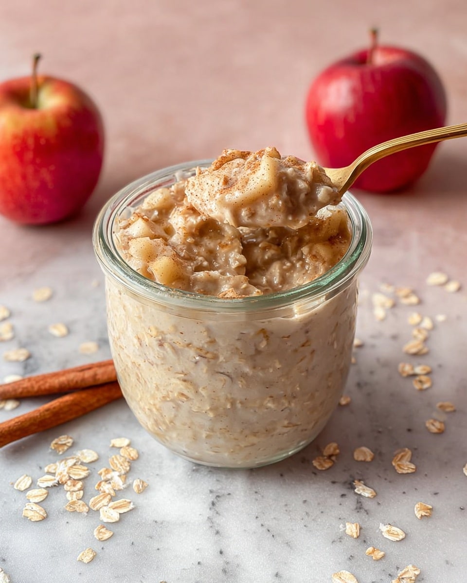 A clear glass jar filled with creamy oatmeal showing a mix of light beige and tan colors, with visible soft oat flakes throughout. A gold spoon scoops up a thick portion of the oatmeal, revealing its smooth, slightly chunky texture. Behind the jar, two red apples sit on a white marbled surface, adding a pop of color, along with some scattered oat flakes and a cinnamon stick lying beside the jar. The background is softly blurred with a white marbled texture, giving focus to the oatmeal in the center. Photo taken with an iphone --ar 4:5 --v 7