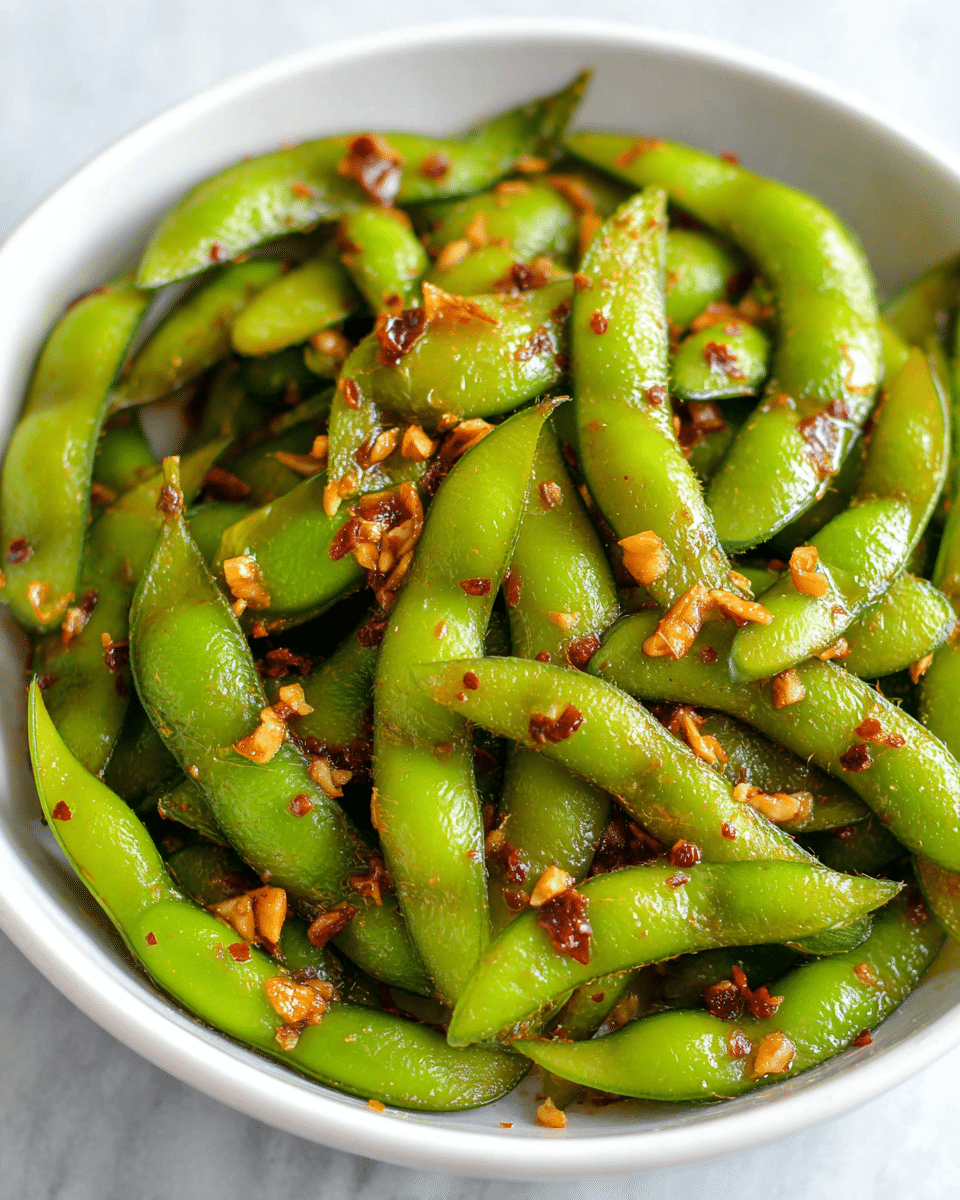 A close-up of a white bowl filled with bright green edamame pods, each coated in a shiny, oily sauce that gives a glistening effect. Small pieces of golden brown garlic and reddish chili flakes are scattered evenly on top and mixed throughout the pods, adding texture and color contrast. The edamame pods appear fresh and slightly fuzzy, nestled closely together. The bowl is placed on a white marbled surface, which adds a clean and light background to the image. Photo taken with an iphone --ar 4:5 --v 7