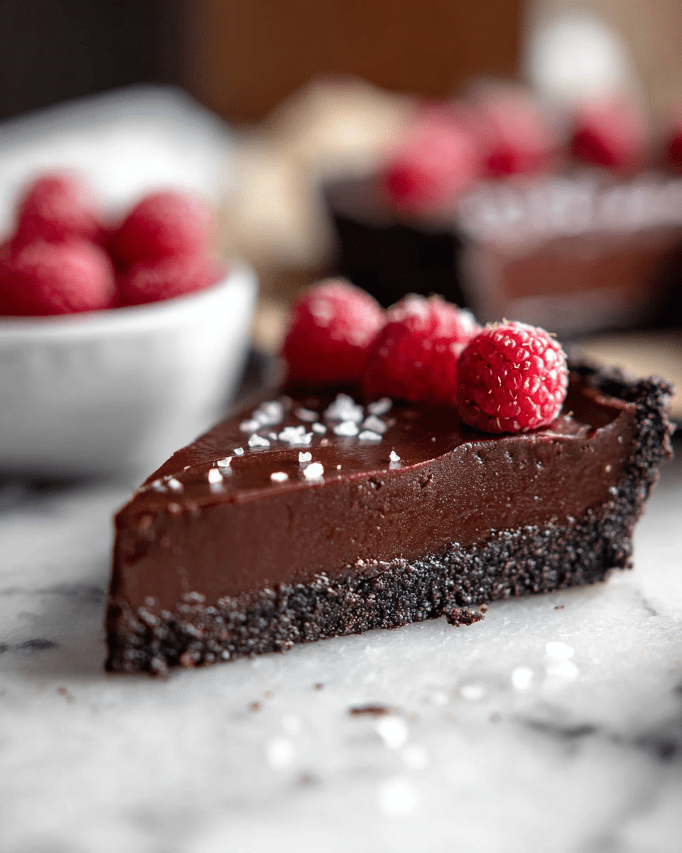 A close-up of a slice of chocolate tart with two visible layers: a dark, crumbly chocolate crust on the bottom and a smooth, thick, glossy chocolate filling on top. The tart slice is decorated with several small, bright red raspberries and a light sprinkling of white salt flakes. In the blurred background, more slices and a white bowl with additional raspberries can be seen, all set on a white marbled surface. photo taken with an iphone --ar 4:5 --v 7