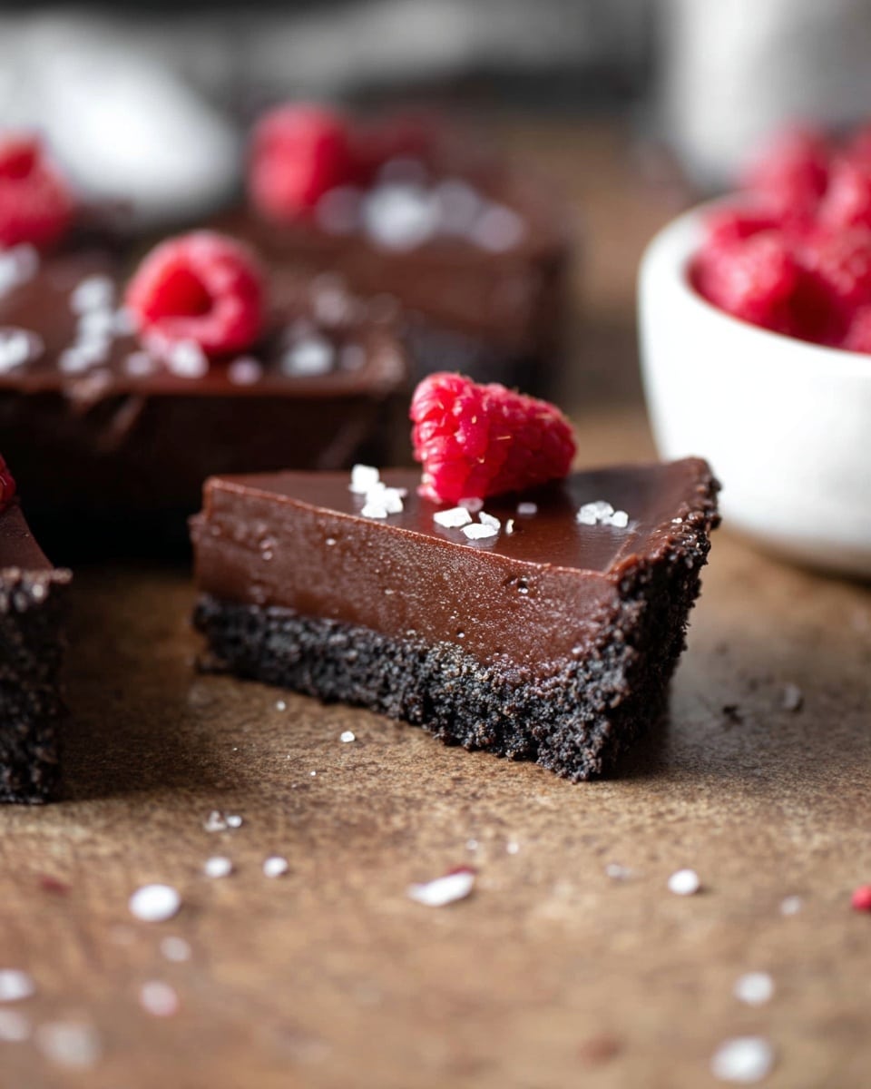 A close-up view of a chocolate dessert with two layers: a dark, crumbly crust at the bottom and a smooth, rich chocolate layer on top. The top is sprinkled lightly with white sea salt flakes and adorned with a few bright red raspberries scattered across the surface. The dessert is cut into a triangular slice and placed on a brown textured surface, with other similar slices blurred in the background and a white bowl containing more raspberries on the right side. The photo taken with an iphone --ar 4:5 --v 7