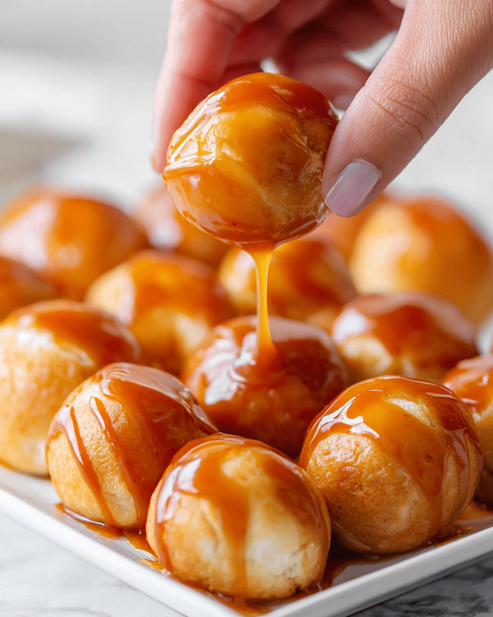 A close-up view of multiple small, round, golden-brown baked dough balls covered in shiny, thick caramel sauce, all placed on a white rectangular dish on a white marbled surface. One dough ball is being held delicately by a woman's hand, with glossy caramel sauce dripping slowly from it. The caramel sauce has a rich amber color with smooth texture, while the dough balls look soft with a lightly cracked surface. Photo taken with an iphone --ar 4:5 --v 7