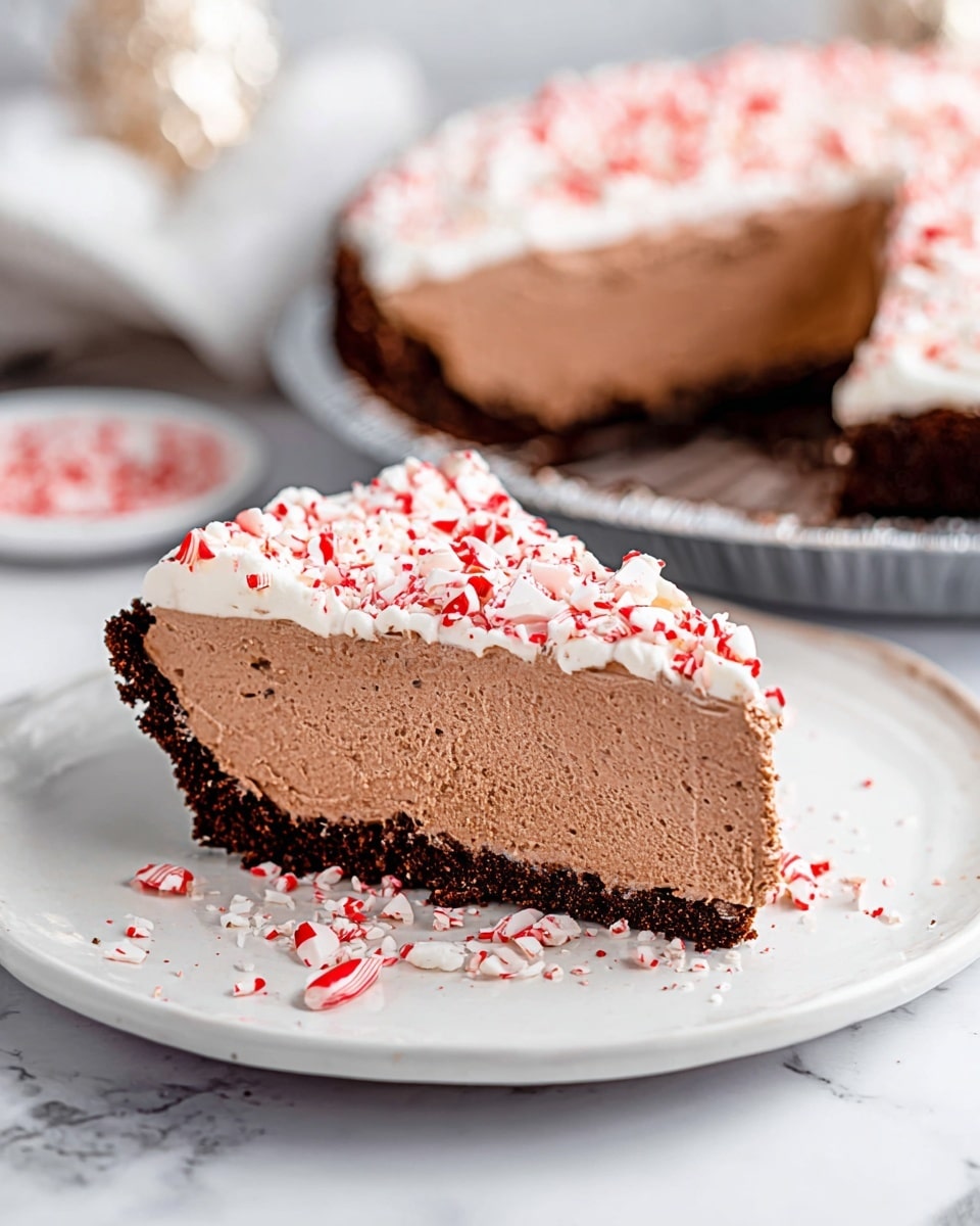 A slice of creamy chocolate mousse pie sits on a white plate with a dark chocolate crumb crust at the bottom, showing one thick layer of light brown mousse with a smooth, airy texture. The top layer is a thin white whipped cream, sprinkled with crushed red and white peppermint candy pieces, scattered around the plate as well. In the background, the rest of the pie is visible in a round metal pan with one slice missing, resting on a white marbled surface. Photo taken with an iphone --ar 4:5 --v 7