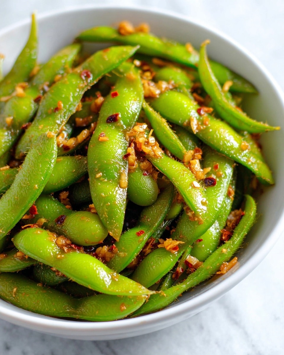 A close-up of green edamame pods covered with small, finely chopped pieces of reddish-brown garlic and spices, all mixed together in a white bowl set on a white marbled surface. The edamame pods are bright green with a shiny, slightly oily texture, and the garlic bits are scattered evenly over the pods, adding a fiery contrast. The bowl is filled to the top, showing the edamame in different directions, some pods overlapping others. photo taken with an iphone --ar 4:5 --v 7