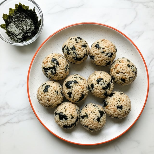 A round white speckled plate with a thin orange rim holds nine rice balls, each coated with dark green seaweed pieces mixed within their light, slightly shiny rice surface; the rice appears sticky and clumped together, with some light brown bits visible inside the balls. In the top left corner, a small clear glass bowl filled with more dark green seaweed sits on a white marbled surface. photo taken with an iphone --ar 4:5 --v 7