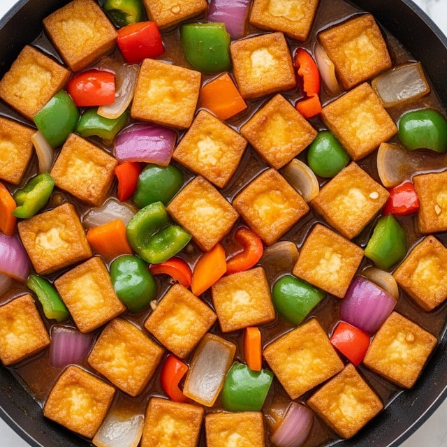 The image shows a close-up of golden brown cubes of tofu mixed with bright, chopped vegetables in a shiny, thick sauce inside a dark pan. The tofu pieces are crisp on the outside and arranged evenly throughout the dish. The vegetables include green, red, and orange bell peppers along with some translucent red onions, all cut into small chunks, adding vibrant colors and texture. The sauce coats all the ingredients, giving a glossy look throughout, and pooling slightly at the bottom of the pan. The background surface around the pan is a white marbled texture. photo taken with an iphone --ar 4:5 --v 7
