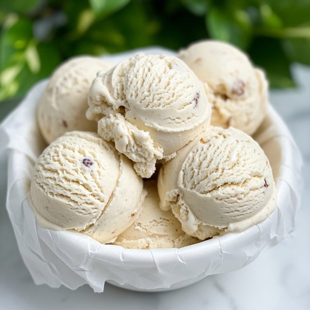 The image shows a white paper-lined bowl filled with several scoops of creamy, off-white ice cream that has small visible bits of nuts or other inclusions inside. The ice cream has a soft and slightly chunky texture, with some smooth folds and rough edges. The background includes green foliage blurred out, and the bowl sits on a white marbled surface. photo taken with an iphone --ar 4:5 --v 7