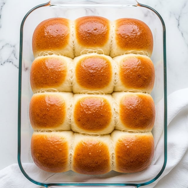 The image shows a clear glass baking dish filled with freshly baked golden brown rolls, arranged closely in two rows of four. Each roll is soft and round with a shiny, smooth top that looks slightly buttery. The rolls have a light, fluffy texture and are touching each other, creating gentle curves where they meet. The dish is set on a white marbled surface with a white cloth underneath, giving a bright and clean look. Photo taken with an iphone --ar 4:5 --v 7