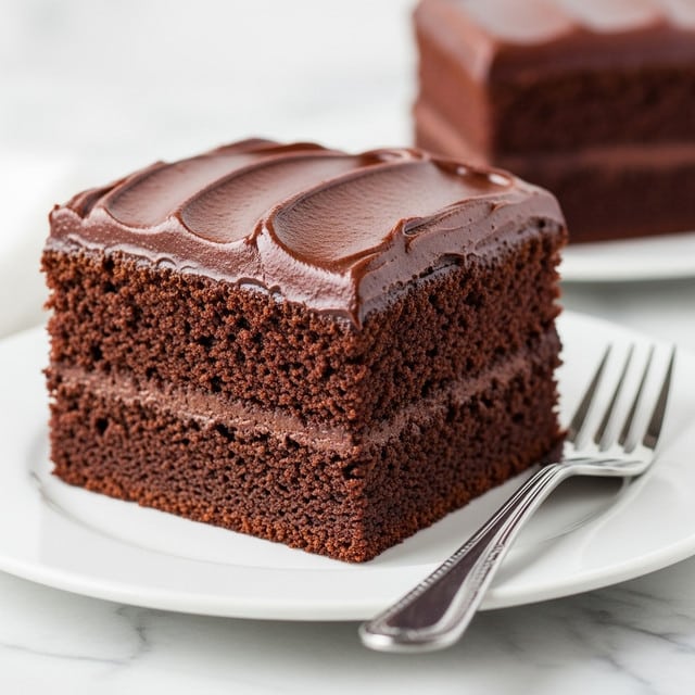 A thick square piece of chocolate cake sits on a white plate with a silver fork next to it on the right side. The cake has two visible layers: a dense, dark brown base with a soft, moist texture, and a smooth, glossy, rich dark brown chocolate frosting layer spread thickly on top with slight swirls and shine. The background is a white marbled texture, and there is a blurred second piece of the same cake in the background. photo taken with an iphone --ar 4:5 --v 7