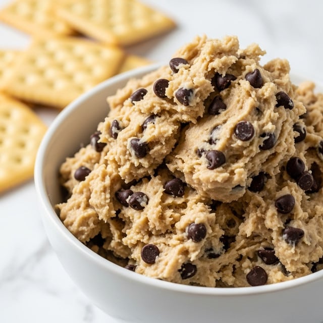 A white bowl filled with a thick, creamy beige dough mixed with many dark chocolate chips, piled high with a chunky, slightly lumpy texture. Behind the bowl, there are a few square crackers that look light and crispy, resting on a white marbled surface. The focus is close on the bowl with soft natural light highlighting the smooth and rich appearance of the chocolate chip cookie dough. photo taken with an iphone --ar 4:5 --v 7