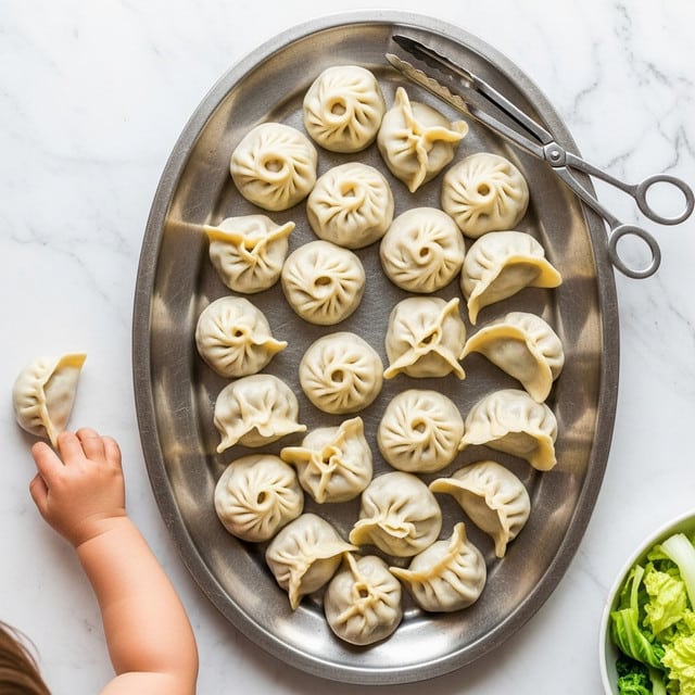 A silver oval platter holds about twenty dumplings, each with a pale beige, slightly shiny dough exterior and soft, rounded shapes, some folded into triangular forms and others with more open tops. The dumplings look smooth and steamed, with subtle creases visible on the dough surface. A pair of silver tongs rests on the upper right edge of the platter. In the lower left corner, a small child's chubby arm and hand reaches to pick up a dumpling. The background is a white marbled texture with a small portion of light green leafy vegetable at the lower right edge. Photo taken with an iphone --ar 4:5 --v 7