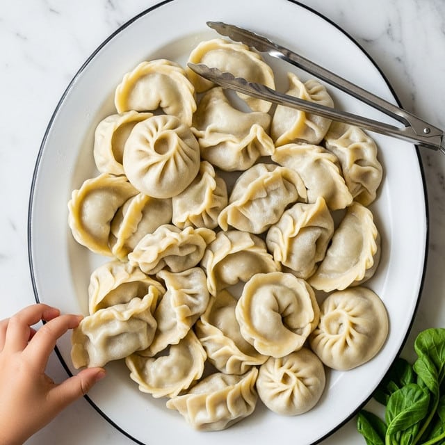 A white oval metal plate holds about 15 cooked dumplings that are light beige with a soft, slightly shiny dough texture, some with folded and pinched edges, others more rounded or flat. The dumplings overlap and fill the plate unevenly. A pair of shiny metal tongs rests on the top right side of the plate, and a small child's hand reaches in from the bottom left, about to pick up a dumpling. The plate is placed on a white marbled surface with some green leafy vegetables visible on the bottom right edge. Photo taken with an iphone --ar 4:5 --v 7