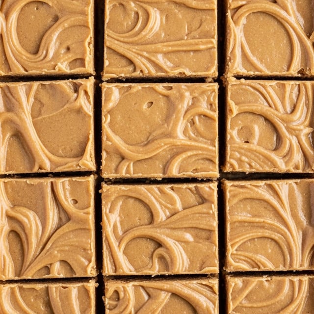 This image shows a close-up of many square pieces of peanut butter fudge arranged tightly side by side. Each piece is light brown with a smooth but slightly grainy texture. The surface of the fudge has soft swirls and small bumps, creating natural patterns across the squares. The fudge looks thick and creamy with clean, straight cut edges that form a grid-like pattern. photo taken with an iphone --ar 4:5 --v 7