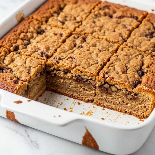 A close-up view of a freshly baked dessert cut into square pieces inside a white ceramic baking dish. The top layer is golden brown with a slightly bumpy and crumbly texture, showing small bits of darker nuts or chocolate throughout. The inside looks soft, moist, and dense with a rich light brown color that contrasts slightly with the crust's darker edges. The dessert fills most of the baking dish, and the edges show some caramelized bits stuck to the white surface. The background is a white marbled texture. photo taken with an iphone --ar 4:5 --v 7