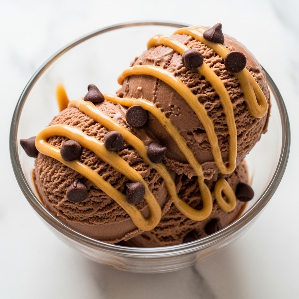 The image shows a close-up view of two scoops of smooth, creamy chocolate ice cream inside a clear glass bowl. Drizzled over the top are thick, golden peanut butter streaks that add contrast and texture. Scattered on the ice cream are several dark, shiny chocolate chips that create a rich and inviting look. The glass bowl sits on a white marbled surface, giving a clean and elegant feel to the presentation. photo taken with an iphone --ar 4:5 --v 7