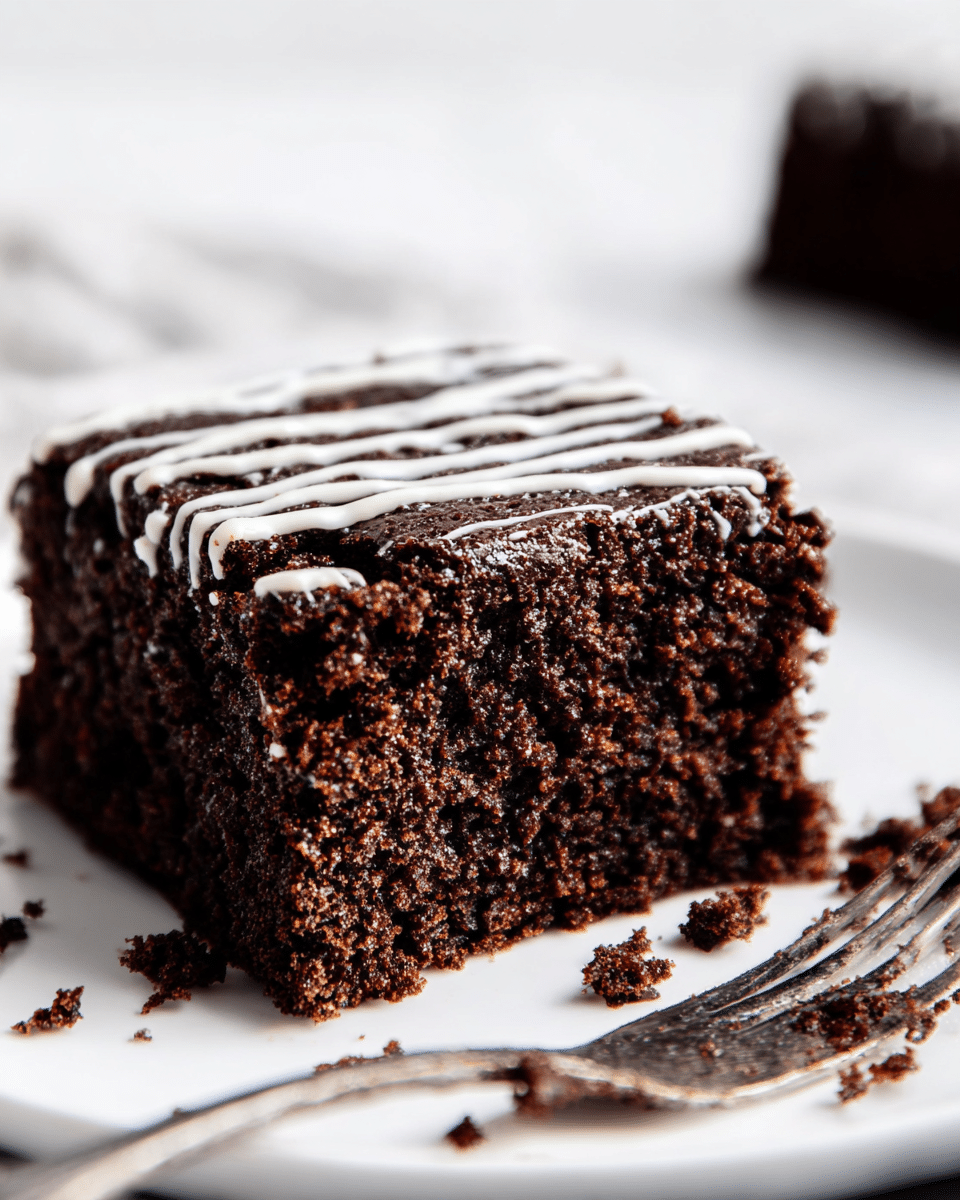A close-up image of a single thick square piece of dark brown chocolate cake with a moist and crumbly texture. The top layer has thin white icing lines drizzled across it in parallel stripes. The cake sits on a white plate with a few cake crumbs scattered around it. In the background, to the right, there is a fork with some chocolate crumbs stuck on it. The whole scene is set on a white marbled surface with soft natural lighting. Photo taken with an iphone --ar 4:5 --v 7