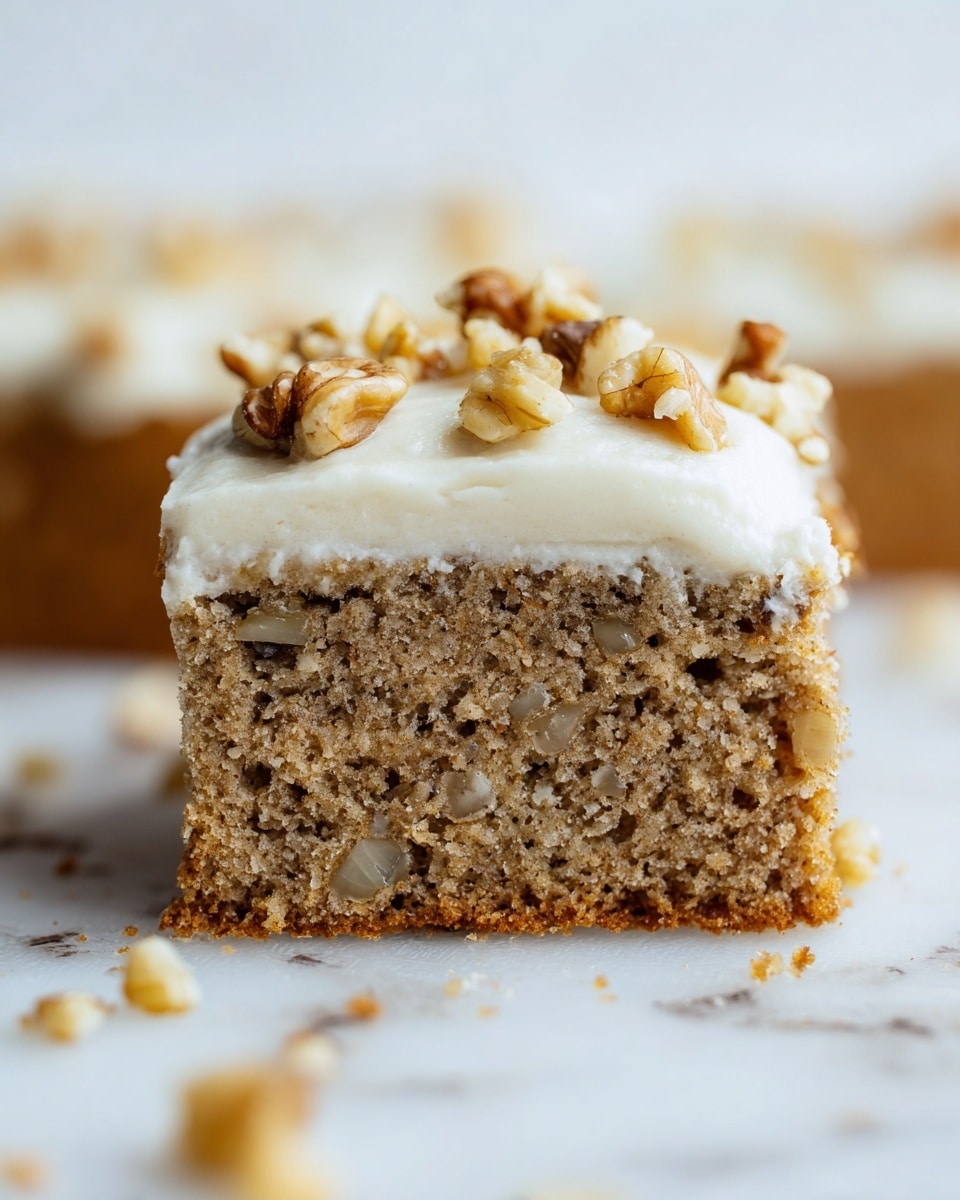 A close-up view of a single square slice of nut bread with two layers: a thick, light brown, moist cake base filled with small pieces of nuts and darker spots, topped with a smooth, creamy white frosting layer that is sprinkled with chopped walnut pieces. The slice is set on a white marbled surface with a soft focus background and some scattered nut crumbs around it. photo taken with an iphone --ar 4:5 --v 7