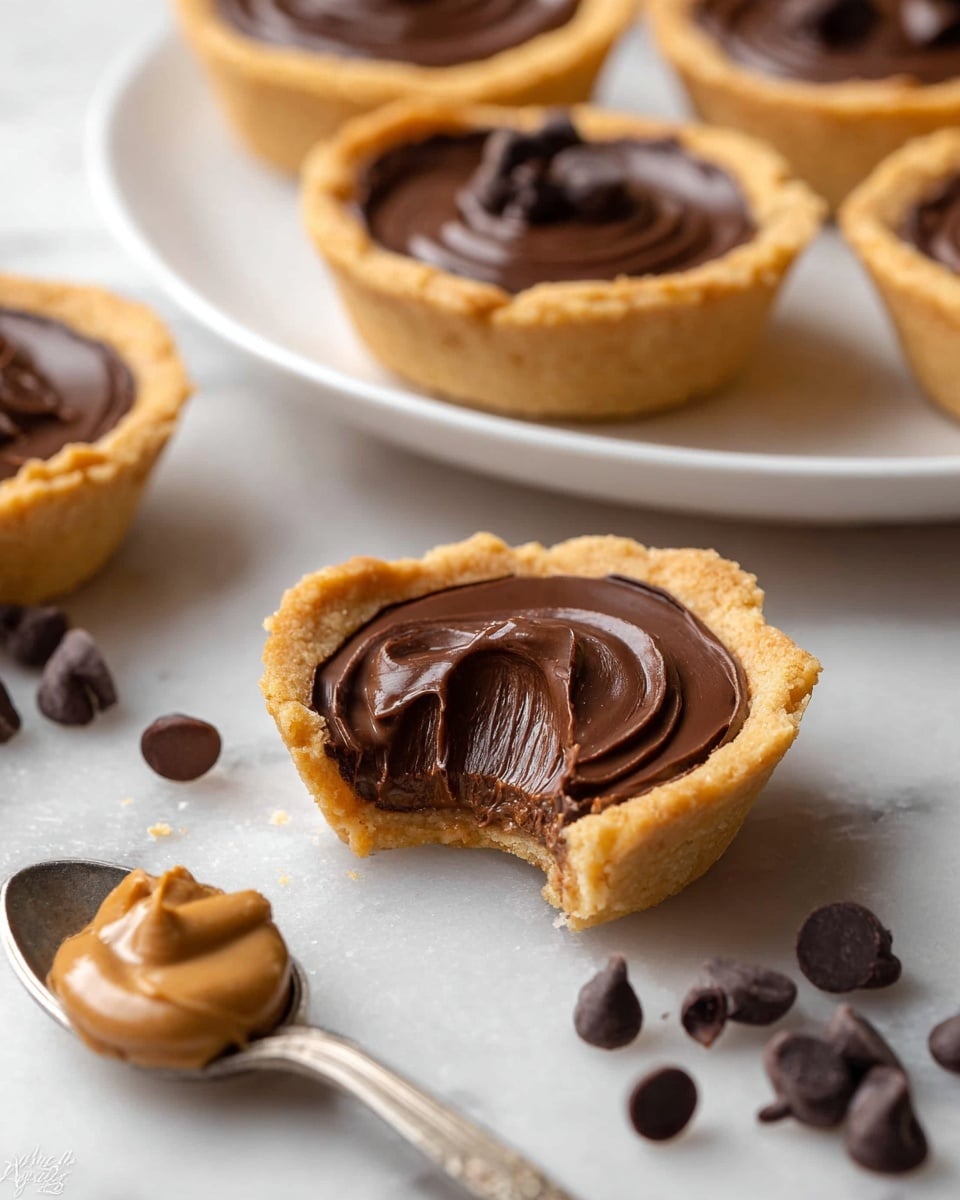 The image shows several small tartlets with a golden cookie crust base and a smooth, thick dark chocolate filling layered inside. One tartlet in the center has a bite taken out, revealing the creamy chocolate filling's dense texture with faint ridges. Around the tarts are scattered dark chocolate chips and a silver spoon with a swirl of light brown peanut butter on it. The tartlets sit on a white marbled surface, with a white plate holding more tartlets blurred in the background. The focus is on the tartlet with the bite taken out, highlighting its glossy, rich chocolate layer photo taken with an iphone --ar 4:5 --v 7