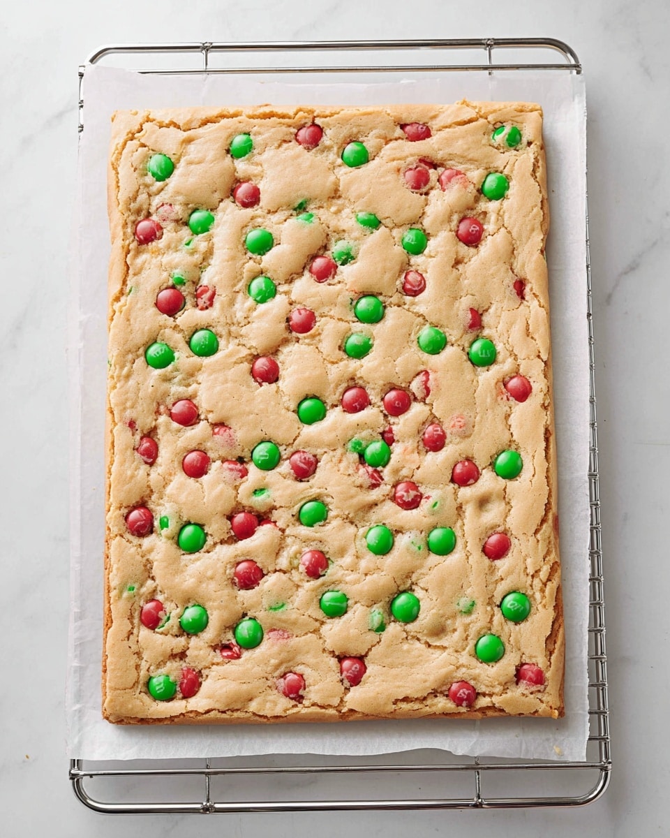 A rectangular baked sheet cookie with a light golden-brown top layer that looks slightly cracked and crisp on the edges. Spread on the surface are scattered red and green candy-coated chocolate pieces, some slightly pressed into the dough and some partially cracked. The cookie rests on white parchment paper, which sits on a metal cooling rack, all placed against a white marbled surface. The cookie is thick and fluffy with a soft texture visible on the edges. photo taken with an iphone --ar 4:5 --v 7