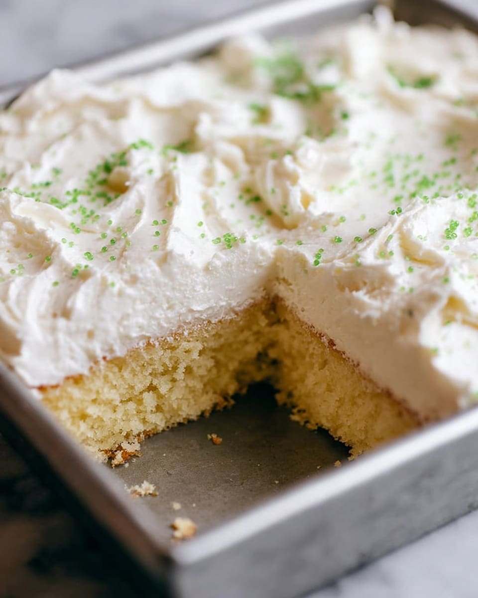 A close-up view of a square cake in a metal pan with one slice removed. The cake has two visible layers: the bottom layer is a soft, light yellow sponge with a moist, crumbly texture, and the top layer is a thick, fluffy white frosting with a textured, swirled surface. Small green sprinkles are scattered lightly across the frosting. The background surface is a white marbled texture. photo taken with an iphone --ar 4:5 --v 7