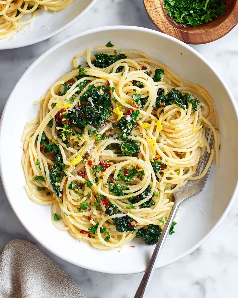 A white bowl filled with spaghetti pasta layered in a light golden-yellow color, mixed with dark green kale leaves spread evenly throughout. Thin slices of garlic and small bits of red chili flakes are scattered over the pasta, with tiny specks of chopped fresh green herbs sprinkled on top. Some bright yellow lemon zest adds contrast, and a silver fork rests inside the bowl on the right side. The bowl sits on a white marbled surface with a wooden bowl of chopped green herbs nearby. Photo taken with an iphone --ar 4:5 --v 7
