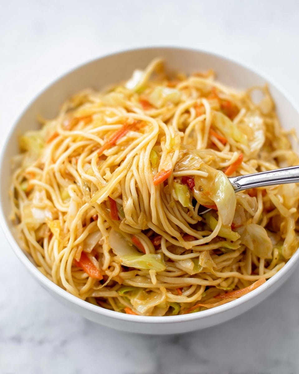 The image shows a white bowl filled with a close-up view of cooked noodles mixed with small pieces of cooked cabbage and bits of carrots, all tossed in a light golden sauce giving a slight shine. The noodles form the main layer, long and thin, light yellow in color with some strands having a slight orange tint from the sauce. The vegetables are scattered throughout, the soft cabbage pieces are pale yellow and the carrot bits provide a bright orange contrast. There is a metal fork inserted into the noodles, lifting some tangled noodles and vegetables from the bowl. The background is a clean white marbled texture. photo taken with an iphone --ar 4:5 --v 7