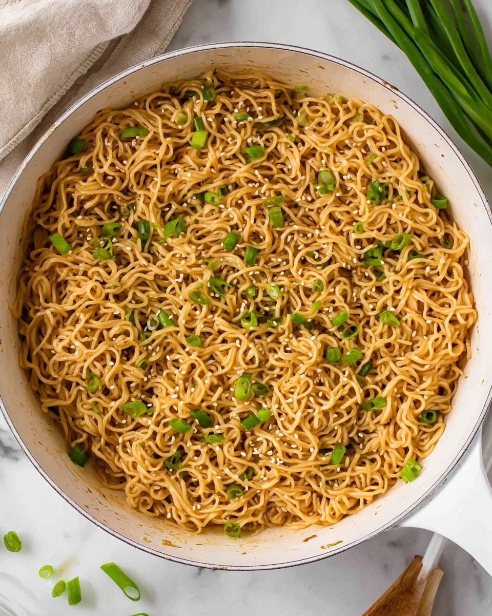 A white pan filled with cooked noodles that are light brown in color, mixed with small pieces of chopped green onions scattered evenly on the top, giving a fresh green contrast. The noodles appear soft and slightly shiny, with a few sesame seeds lightly sprinkled, adding texture. The pan sits on a white marbled surface, with some green onion pieces and a white spatula partially visible around it. photo taken with an iphone --ar 4:5 --v 7