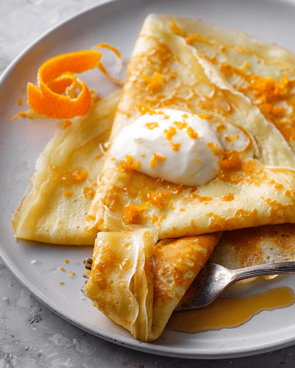 Four thin crepes folded into triangles are placed in a round white plate, overlapping each other. The crepes are light yellow with golden-brown spots and are sprinkled with orange zest and drizzled with honey or syrup, making them shiny. In the center on top of the crepes, there is a dollop of white whipped cream with a little orange zest on it. Two small, decorative orange peel spirals are placed on opposite sides of the plate. The plate is set on a white marbled surface with a textured gray cloth partially visible at the bottom left. photo taken with an iphone --ar 4:5 --v 7